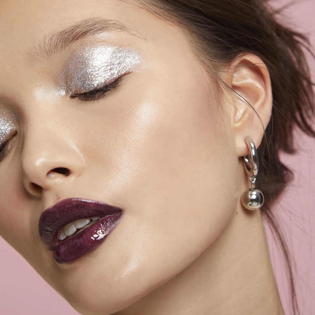 Close-up of a woman's face with silver eye makeup and purple lipstick, wearing a silver hoop earring on a pink background.