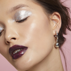 Close-up of a woman's face with silver eye makeup and purple lipstick, wearing a silver hoop earring on a pink background.