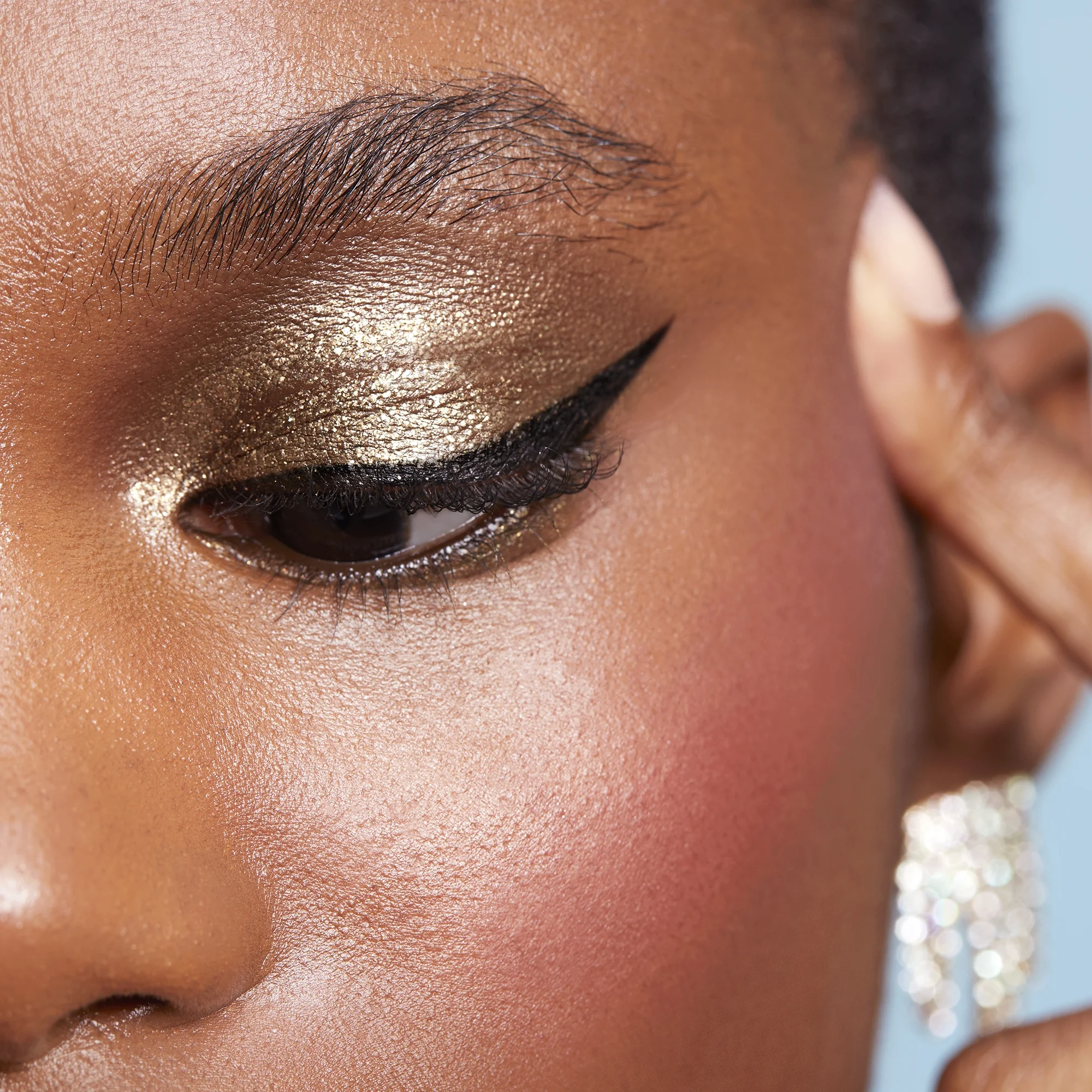 Close-up of a woman's eye with gold eyeshadow and black eyeliner, wearing a pearl earring.
