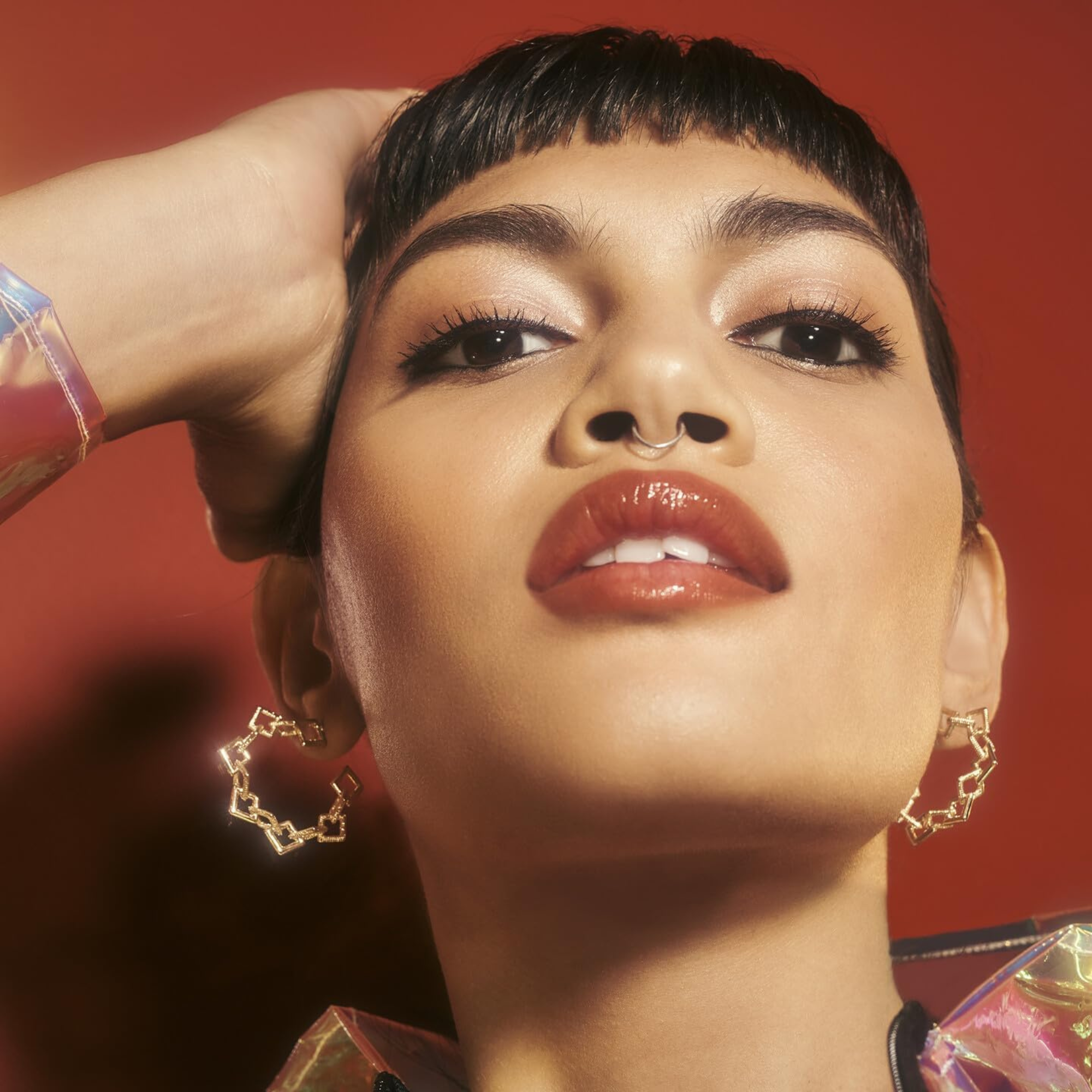 Close-up of a woman with gold hoop earrings against a red background wearing elf sheer lipstick